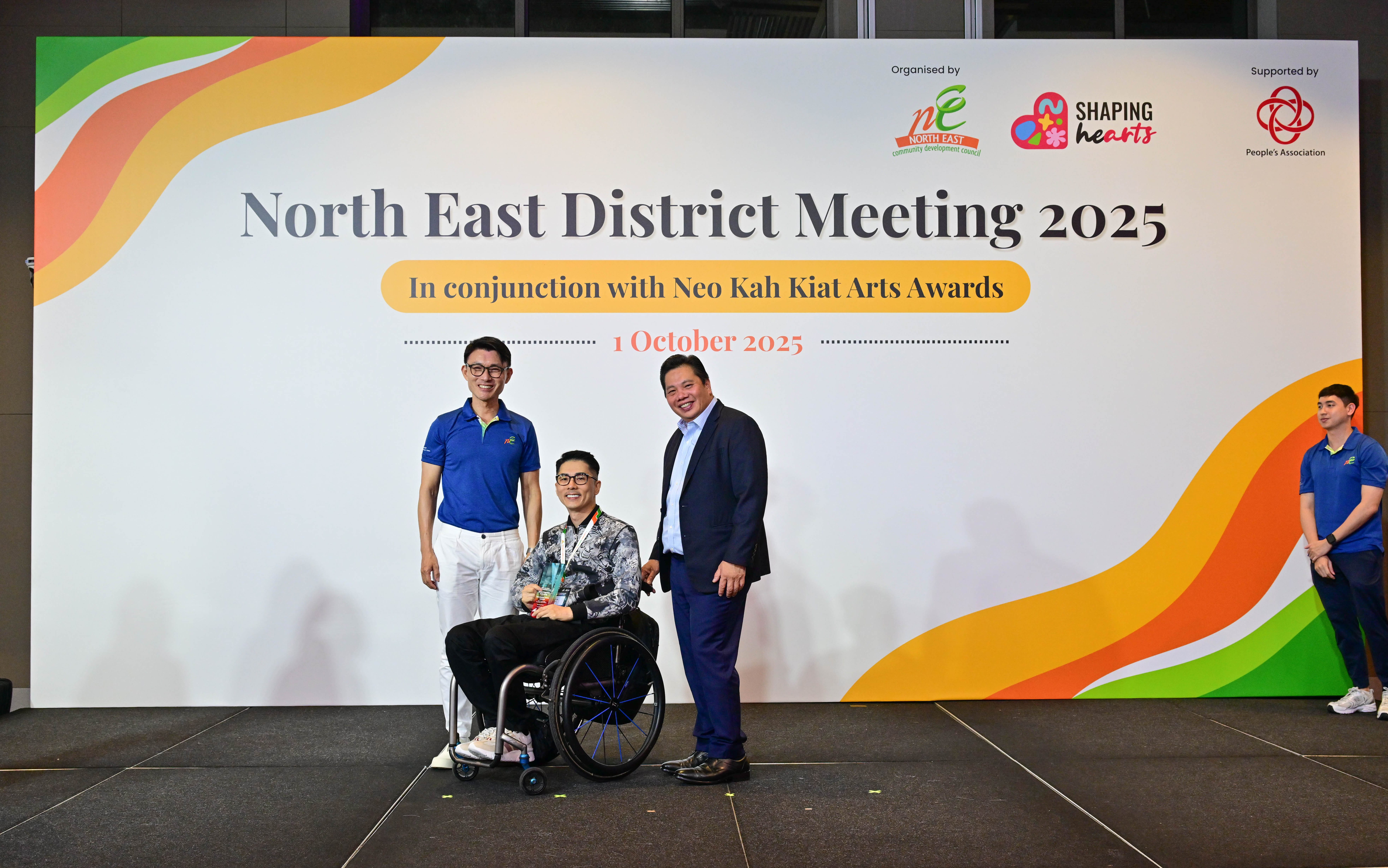 Award recipient standing on stage with the presenter, holding a acrylic plaque and posing for a group photo during the award ceremony, with a backdrop reading ‘North East District Meeting 2025 In conjunction with Neo Kah Kiat Arts Awards'
